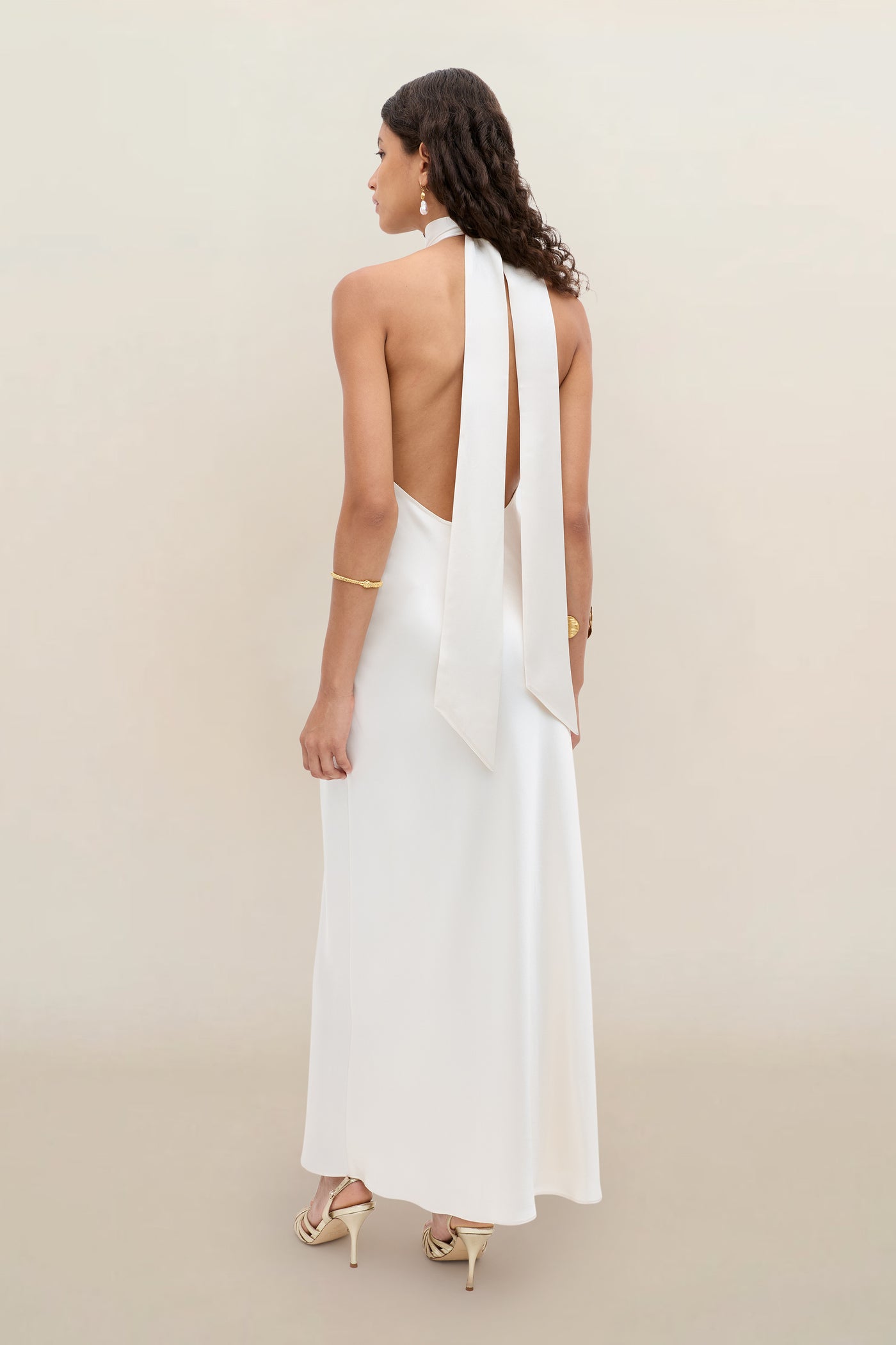 A woman with wavy dark hair models the Arista - Pearl, an elegant white halter dress with an open back and long ties, paired with gold jewelry and heeled sandals against a neutral beige background.