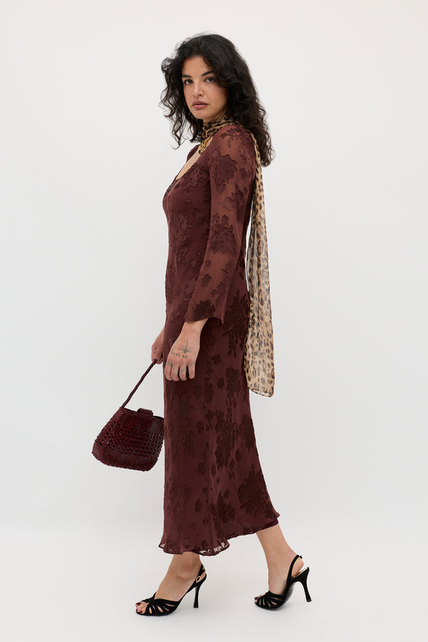A woman with curly dark hair wears the Noraya - Bouquet Devoré Brown dress, paired with black heels, a beige printed scarf, and carries a dark red woven handbag. She stands against a plain white background.
