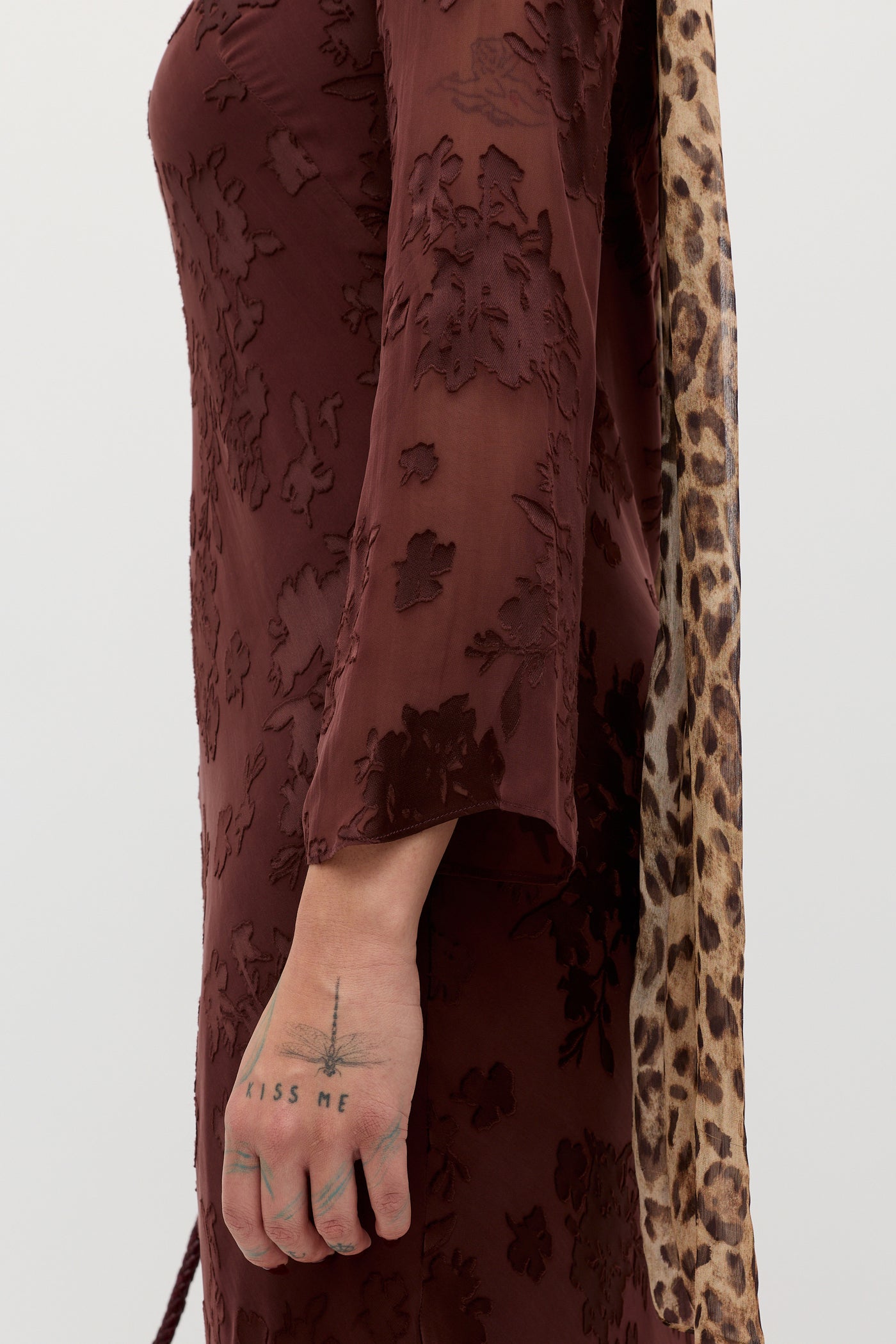 A person wears a textured maroon dress and lets their arm hang down. The Noraya - Bouquet Devoré Brown leopard-print scarf hangs from their shoulder. On their hand is a "KISS ME" tattoo with a small line drawing above it.