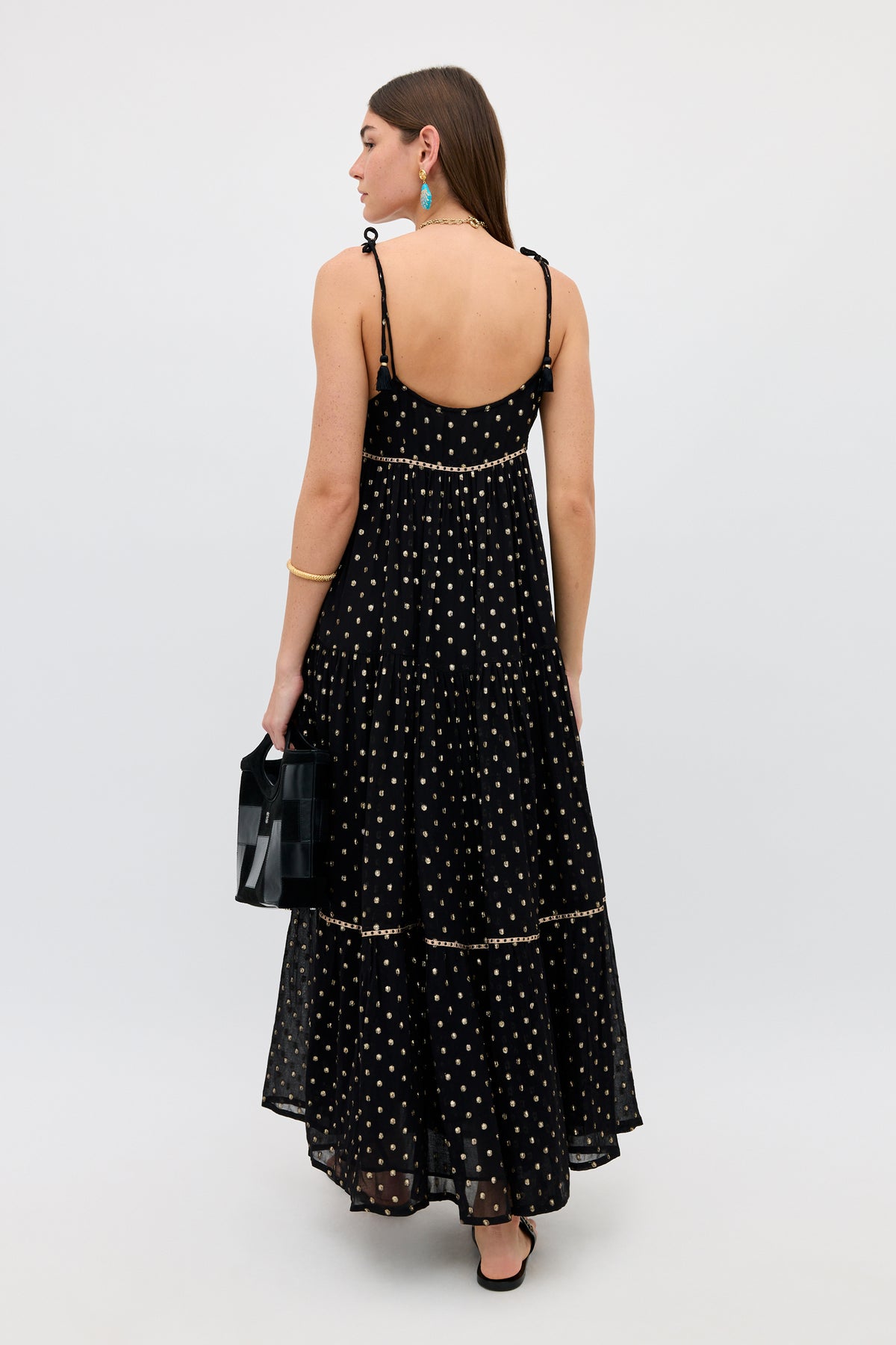 A woman stands facing away, wearing the Kamilah - Black sleeveless maxi dress with white polka dots and holding a small black handbag. She has long brown hair, blue earrings, and black sandals. The background is plain and light-colored.