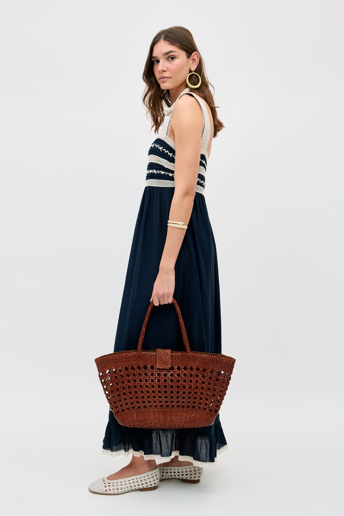 A woman with long brown hair in a sleeveless navy and white maxi dress holds the Lucine - Tan bag, crafted from woven leather. She wears gold hoop earrings, a gold bracelet, and white woven shoes against a plain light background.