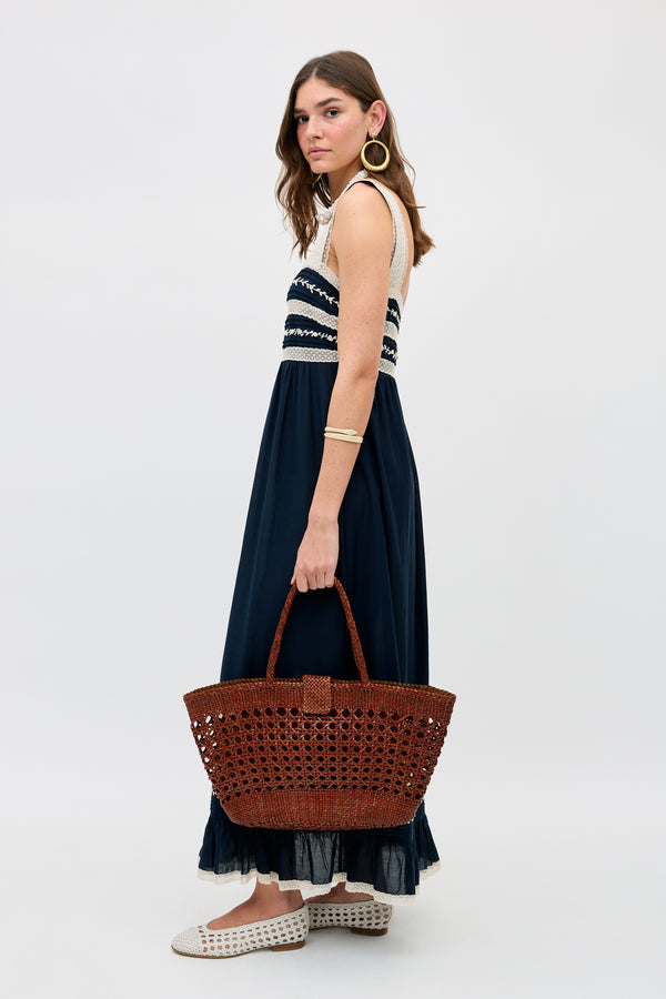 A woman with long brown hair in a sleeveless navy and white maxi dress holds the Lucine - Tan bag, crafted from woven leather. She wears gold hoop earrings, a gold bracelet, and white woven shoes against a plain light background.