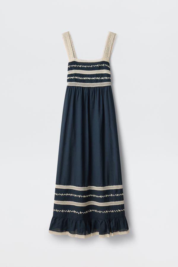 The Malie - Deep Navy is a sleeveless maxi dress featuring wide cream lace-trimmed straps, cream and white embroidered stripes on the bodice and hem, and a ruffled bottom edge.