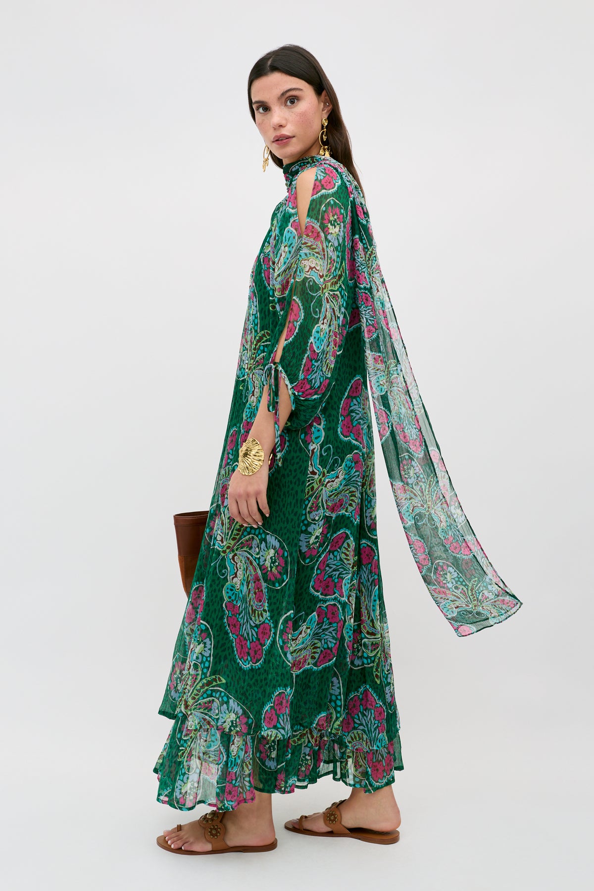 A woman wears the Hayden - Butterfly Bloom Green dress featuring a long, floral design with pink and yellow accents, open shoulders, and a flowing scarf detail. She pairs it with a brown bag, flat sandals, and gold jewelry against a light background.