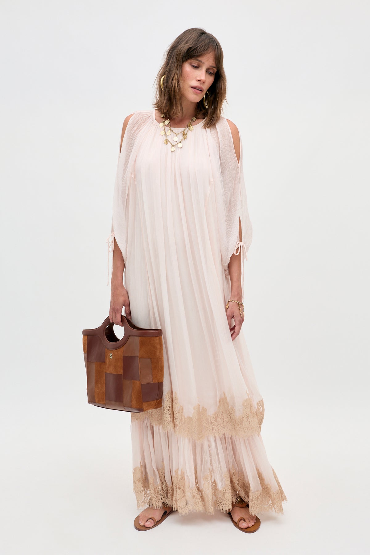 A woman wearing the Hayden - Shell, a flowing light pink dress with lace trim, holds a brown patchwork bag and wears sandals as she stands against a plain white background.