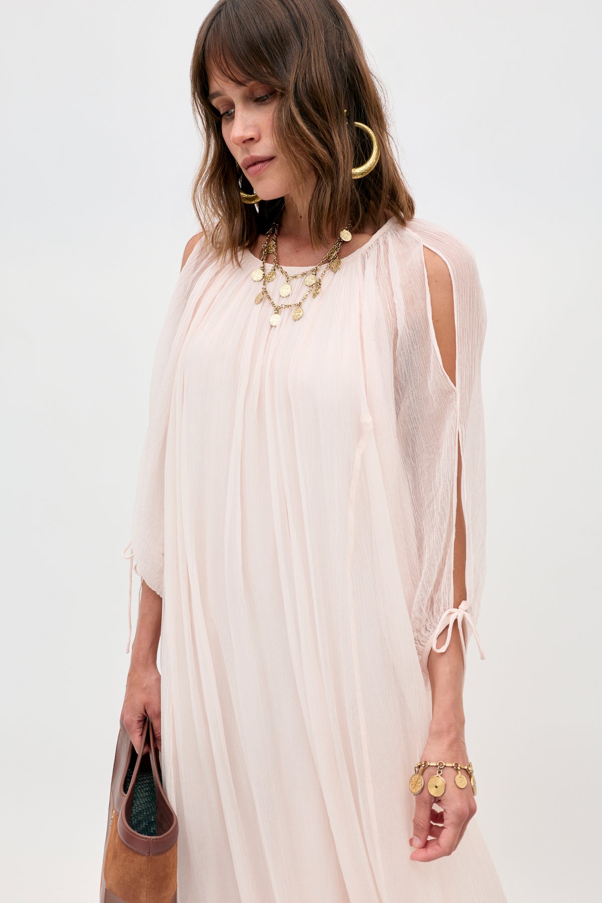 A woman wearing the Hayden - Shell light, flowy blush pink dress with shoulder cutouts, styled with gold hoop earrings, a layered necklace, and bracelet, holds a brown clutch against a plain background.