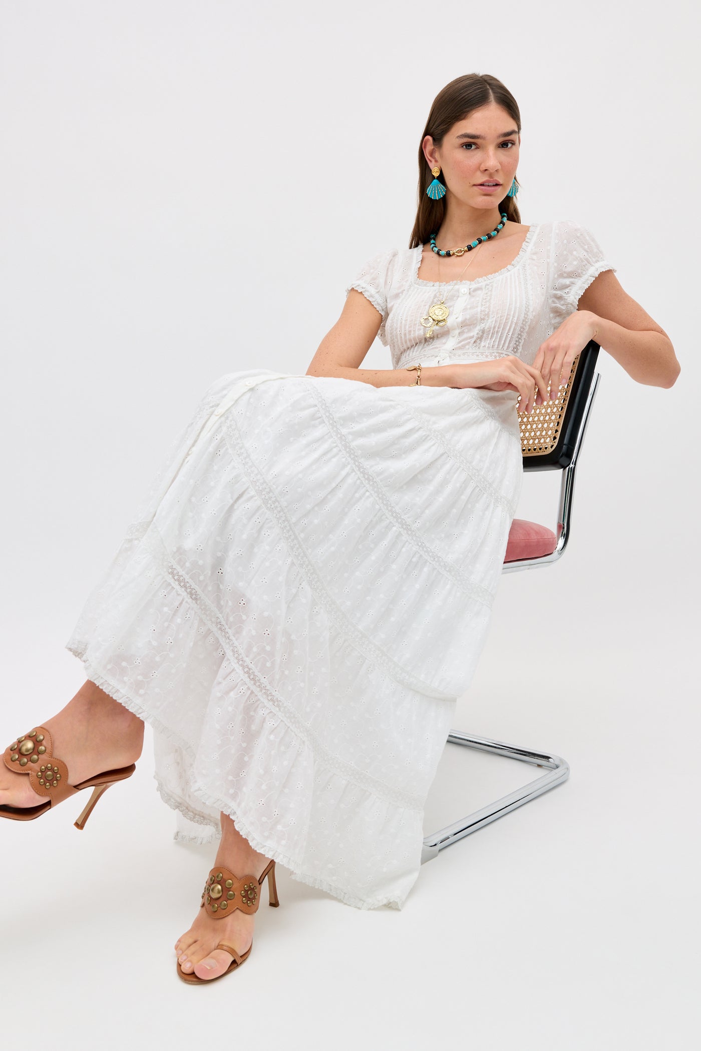A woman wearing the Jaclyn - Ivory long white broderie anglaise summer dress, beige heeled sandals, large turquoise earrings, and layered necklaces sits on a modern pink and metal chair against a plain white background.