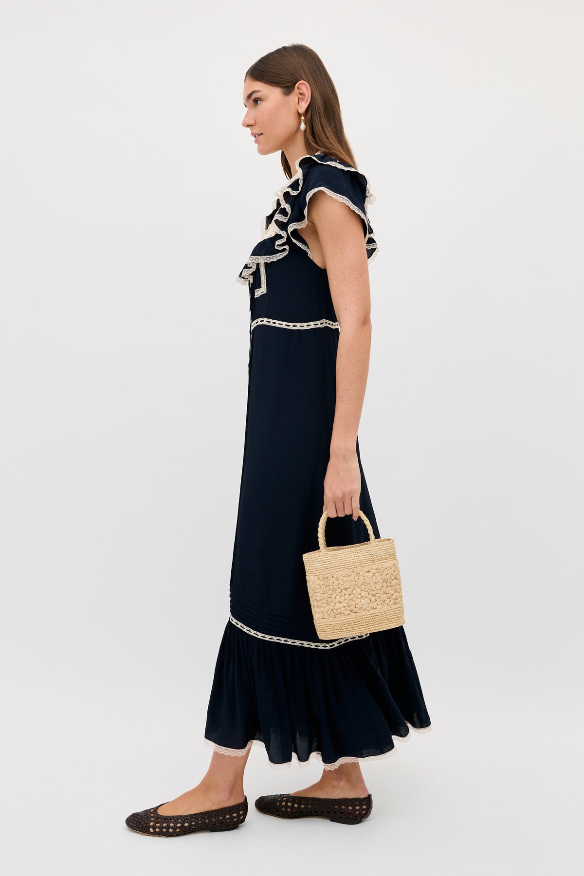 A woman stands in profile wearing the Kendara - Deep Navy cotton midi dress with ruffled details and white trim, holding a woven straw handbag. She pairs it with black woven flats and drop earrings against a plain light background.