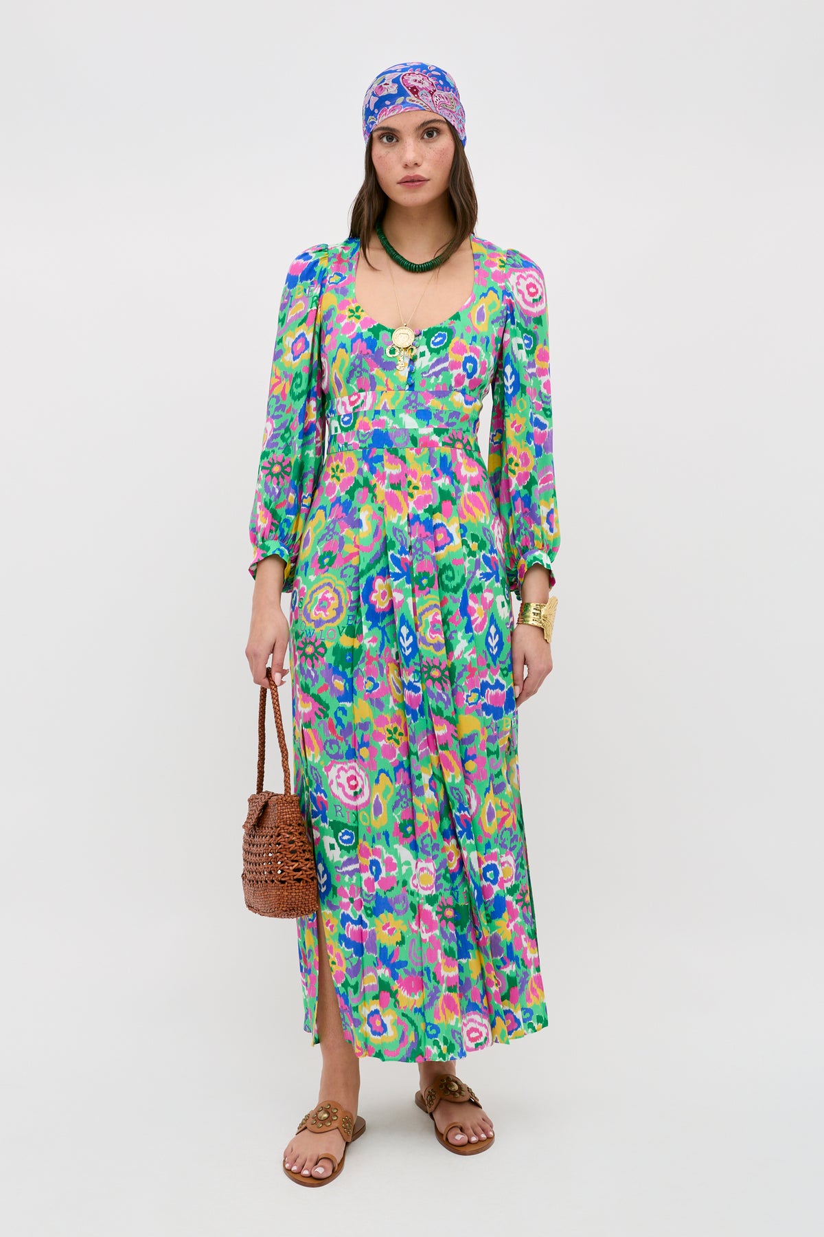 A woman in the "Alfie - Ikat Bloom Green" long floral dress, with a matching headscarf, brown sandals, woven bag, and gold jewelry stands against a white background.