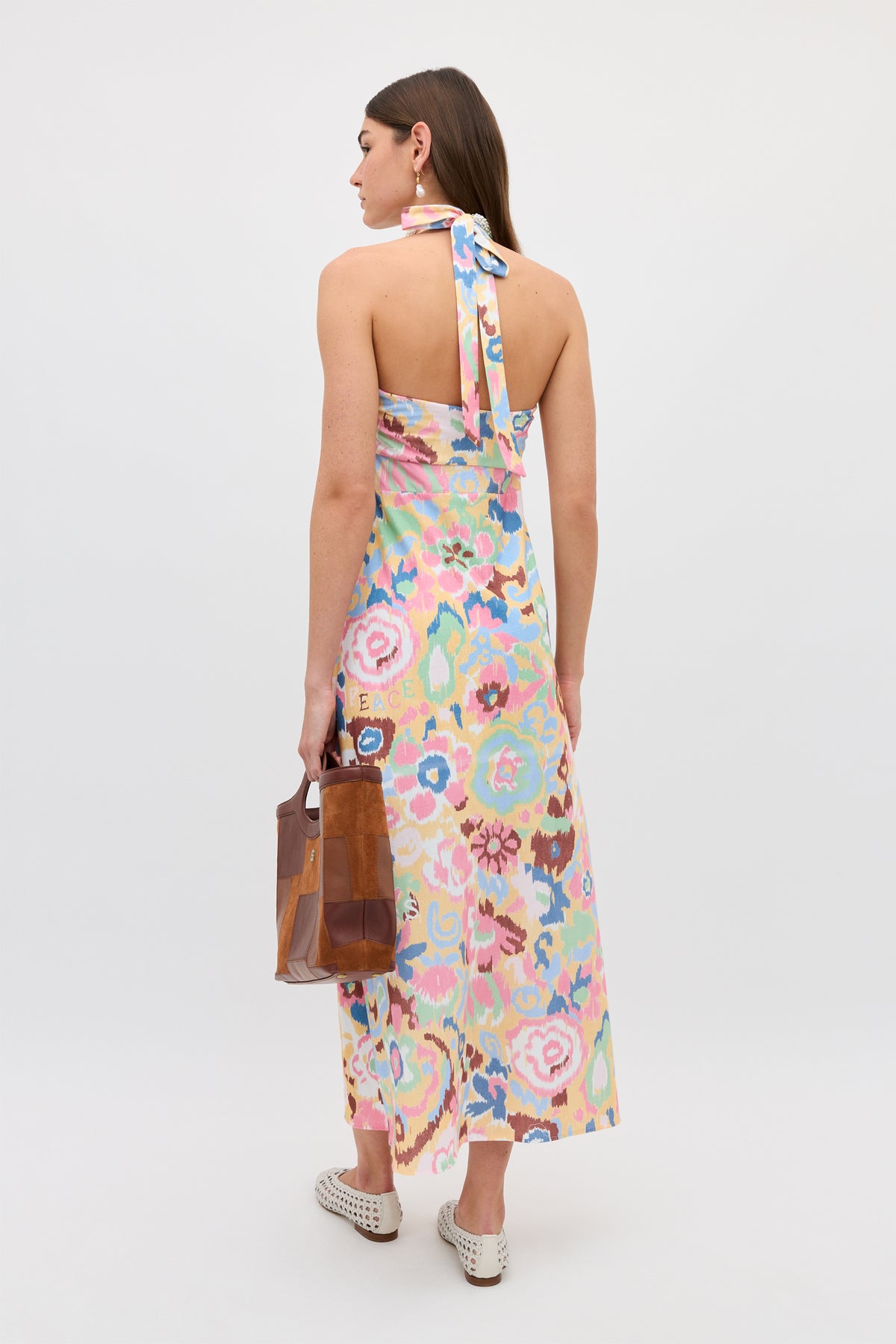 A woman faces away, wearing the Ilyana - Ikat Bloom Yellow floral halter dress. She holds a brown patchwork handbag and wears white woven slip-on shoes, all set against a plain white background.