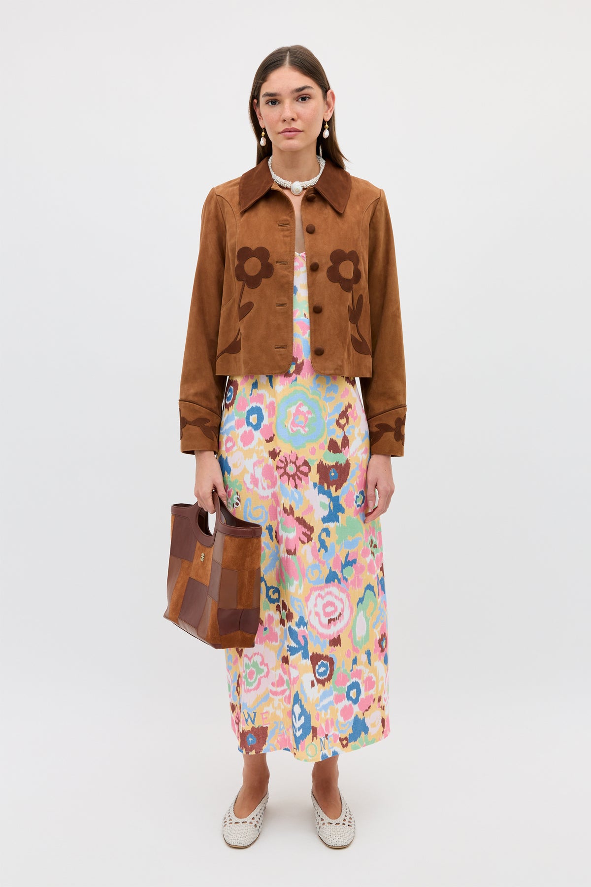 Against a plain background, a woman wears the Ilyana - Ikat Bloom Yellow halterneck dress, styled with a brown suede jacket, patchwork handbag, white slip-on shoes, and a beaded necklace for an elegant look.