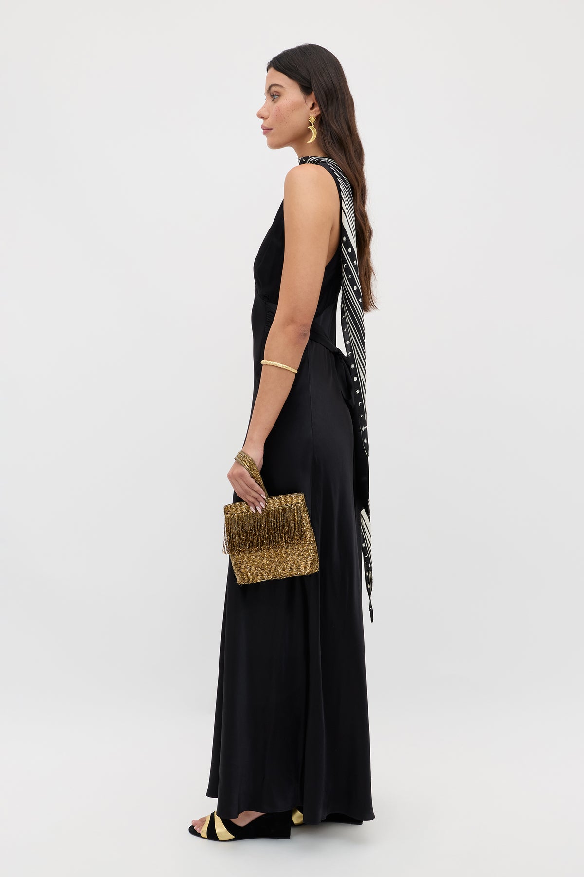 A woman stands in profile wearing the Evalise - Black dress against a plain white background. She accessorizes with a gold clutch, black open-toe heels, gold jewelry, and a black-and-white scarf draped over her shoulder.