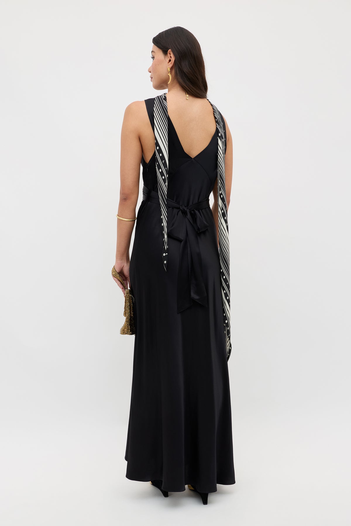 A woman stands facing away, wearing the Evalise - Black long sleeveless dress with a v-shaped back and tied waist. She drapes a black-and-white scarf over her shoulders, holds a small clutch, and accessorizes with gold jewelry.