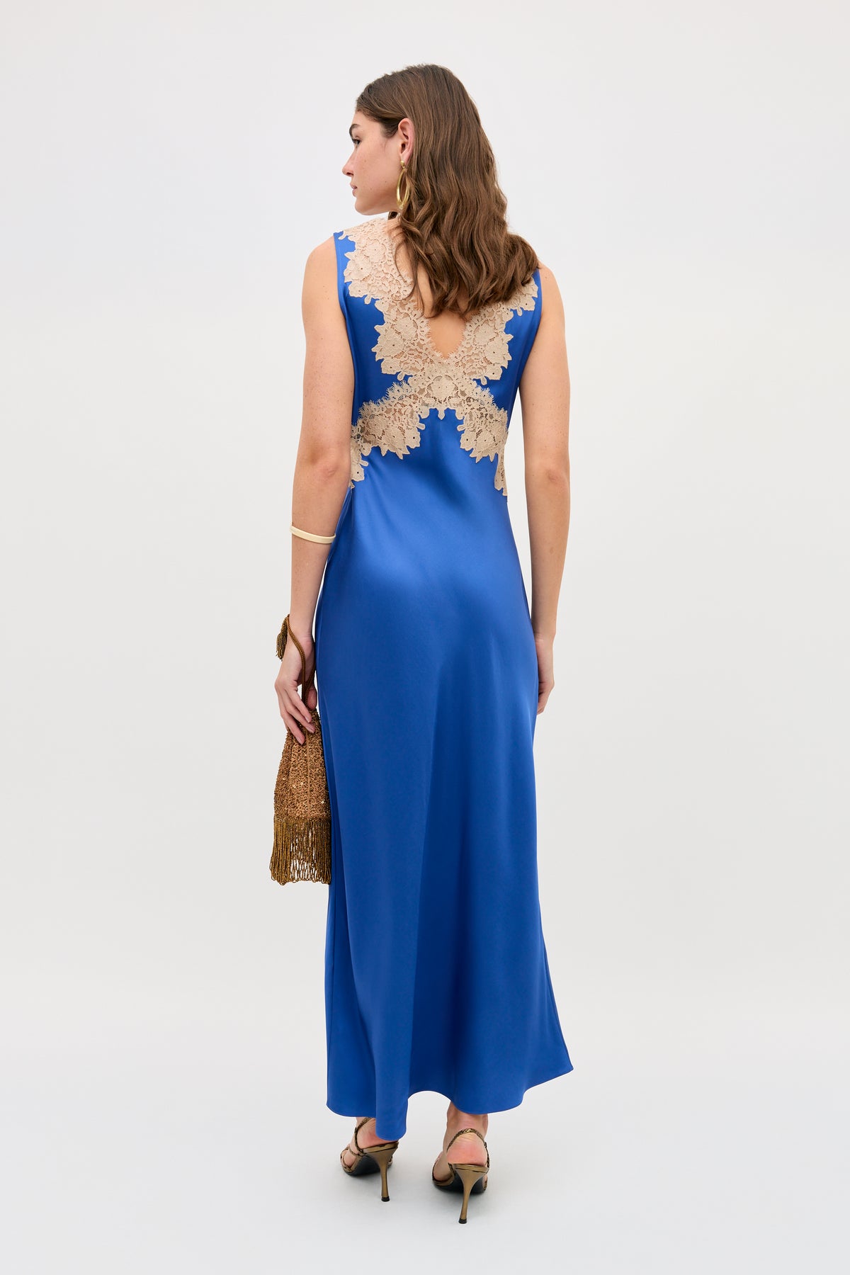 A woman stands in heels against a plain light background, wearing the Lysandra - Iris Blue sleeveless floor-length gown featuring beige lace back detailing and holding a gold clutch.