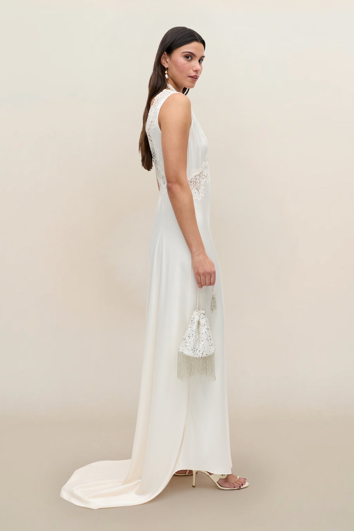 A woman in the Azzurra - Pearl sleeveless, floor-length lace wedding dress holds a small beaded handbag. She has long brown hair, wears high heels, and poses against a neutral background.