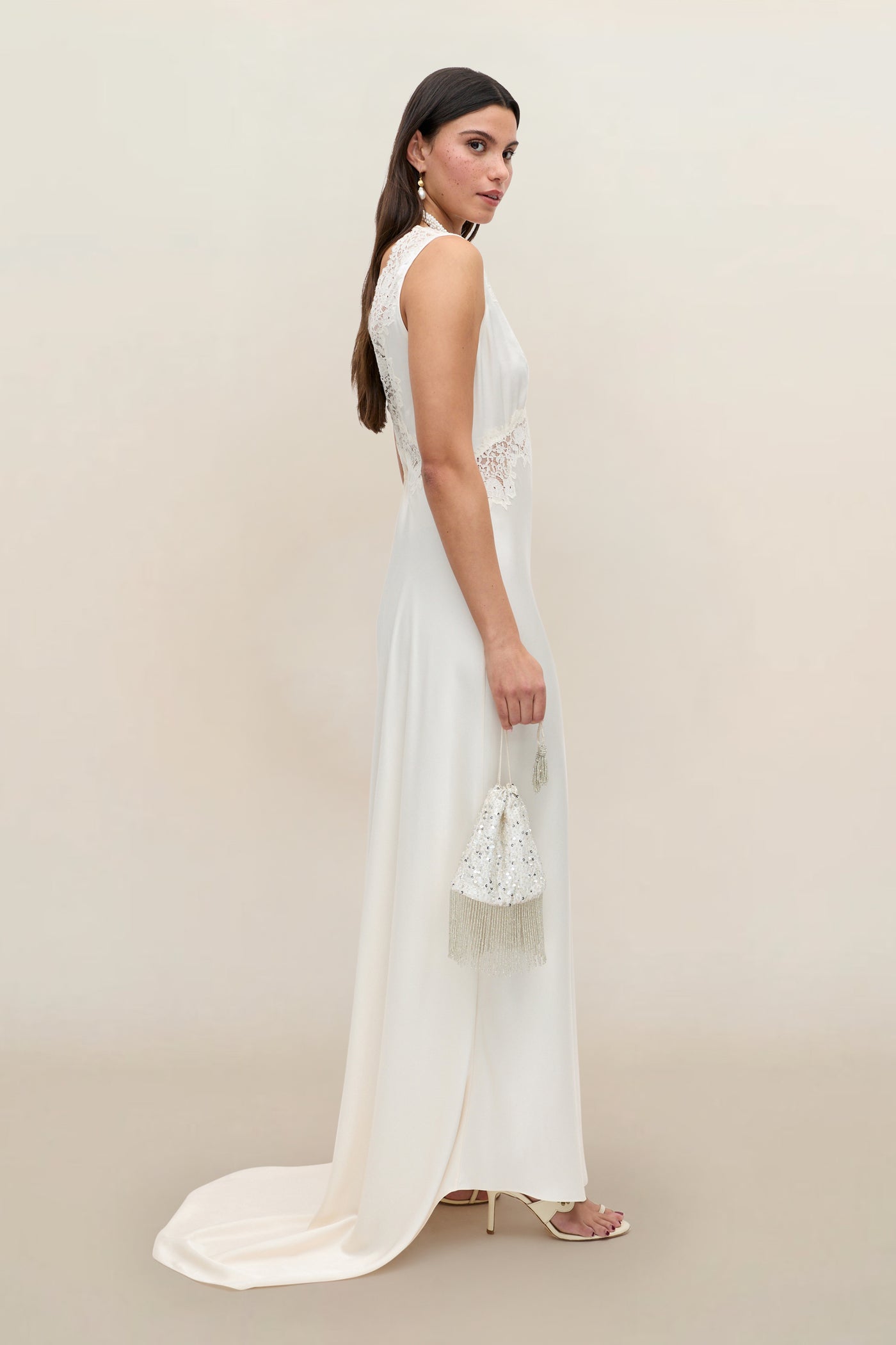 A woman in the Azzurra - Pearl sleeveless, floor-length lace wedding dress holds a small beaded handbag. She has long brown hair, wears high heels, and poses against a neutral background.