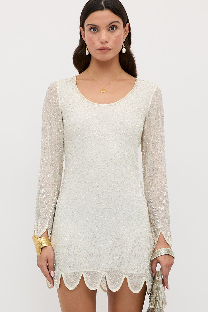 A woman stands against a white background wearing the Dulce Florence Embellishment Pearl mini dress with long sleeves, scalloped hem, styled with pearl drop earrings, a gold necklace and bracelet, and holding a small beaded bag.