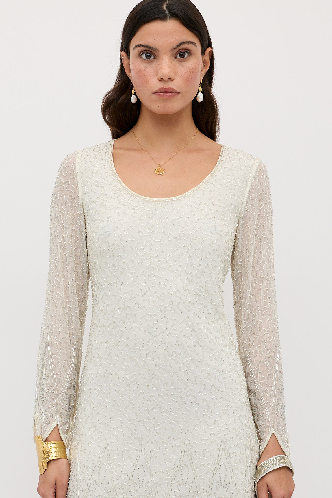 A woman with long brown hair wears the Dulce – Florence Embellishment Pearl dress, featuring cream-colored textured fabric and sheer sleeves, styled with gold drop earrings, standing against a plain light background.