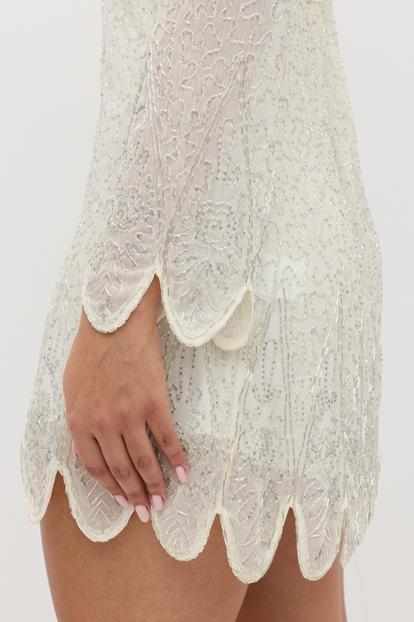 A close-up shows a person wearing the Dulce - Florence Embellishment Pearl dress, featuring intricate beading and lace on white long sleeves with scalloped edges. Their hand rests by their side.