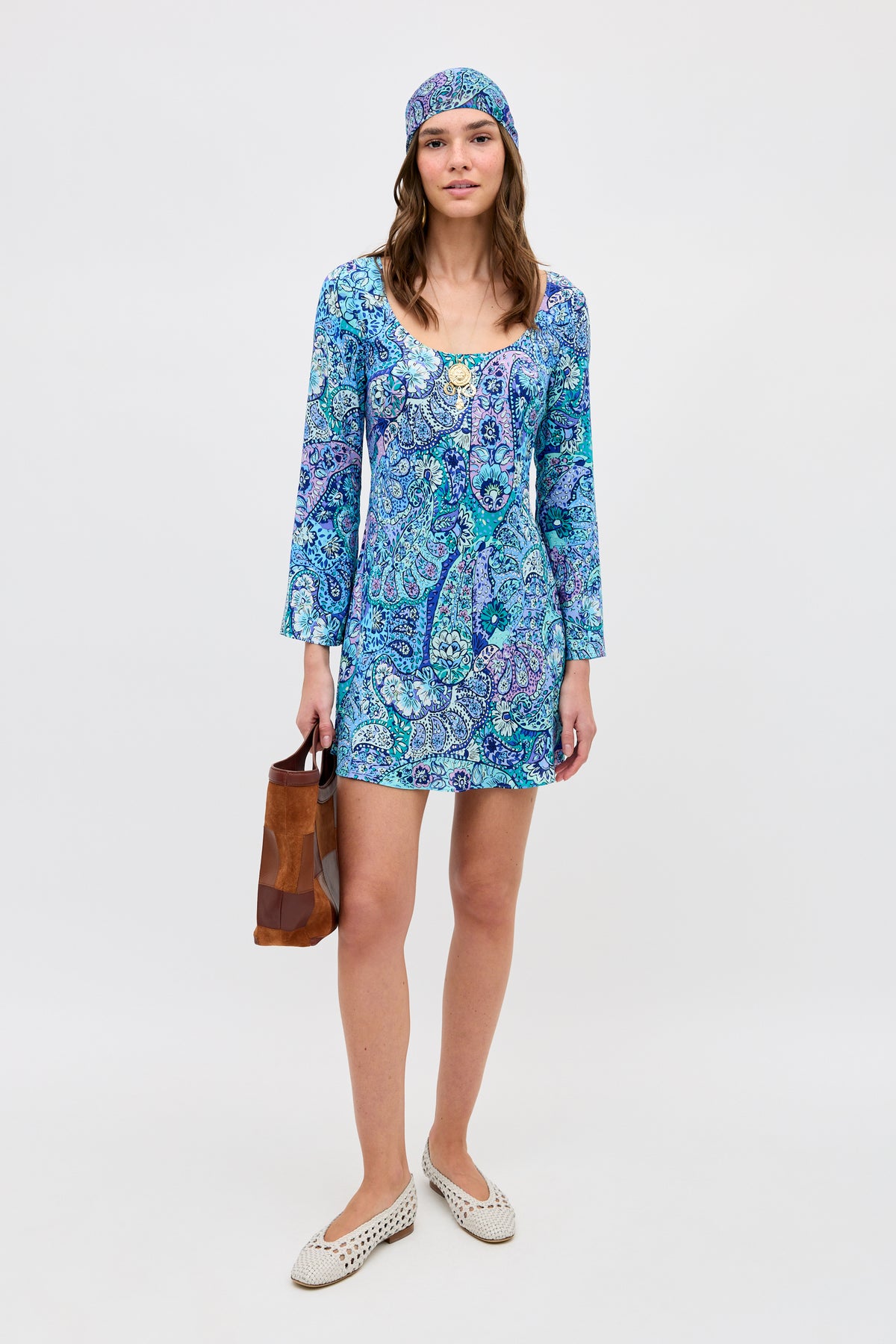 A woman stands against a plain white background, wearing the Dulce - Paisley Floral Blue dress with a matching headscarf. She is holding a brown bag, has on white slip-on shoes, and appears relaxed.