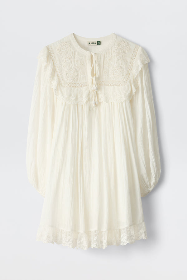 The Inez - Lilly Embroidery Ivory is a loose-fitting, long-sleeved ivory dress with lace details on the yoke, cuffs, and hem. It features a tie neckline and a softly gathered, flowy silhouette.