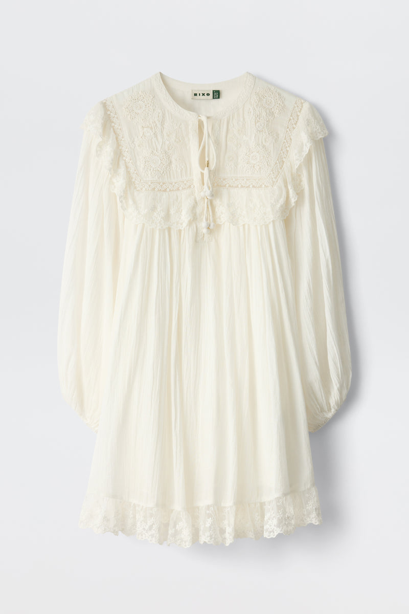 The Inez - Lilly Embroidery Ivory is a loose-fitting, long-sleeved ivory dress with lace details on the yoke, cuffs, and hem. It features a tie neckline and a softly gathered, flowy silhouette.