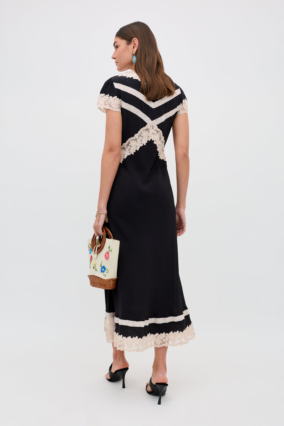 A woman faces away, wearing the Martina - Black midi dress with cream lace trim and X-shaped back detail. She carries a white floral embroidered handbag, black heeled sandals, and turquoise earrings.