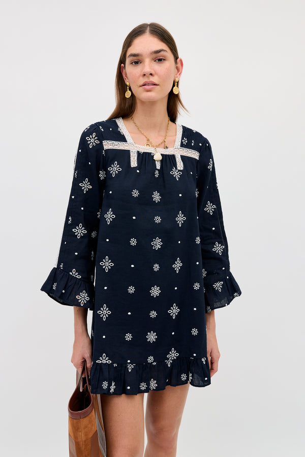 A person wears the Susana - Blossom Cutwork Embroidery Navy dress, featuring navy linen-cotton fabric adorned with white floral patterns, ruffled sleeves and hem, and delicate white lace detailing. They accessorize with a brown bag and gold jewelry while standing against a plain light background.