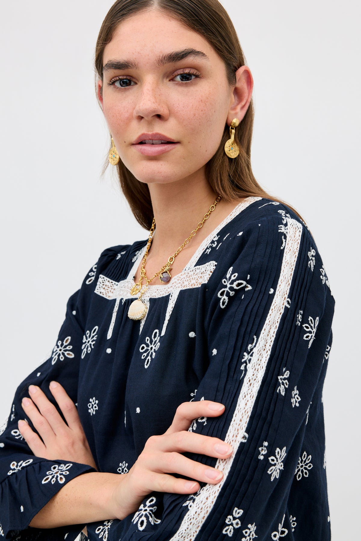 A woman with long brown hair wears the Susana - Blossom Cutwork Embroidery Navy blouse, featuring elegant white lace detailing and intricate embroidery. She poses with her arms crossed, accessorized with gold earrings and a necklace, against a plain light background.
