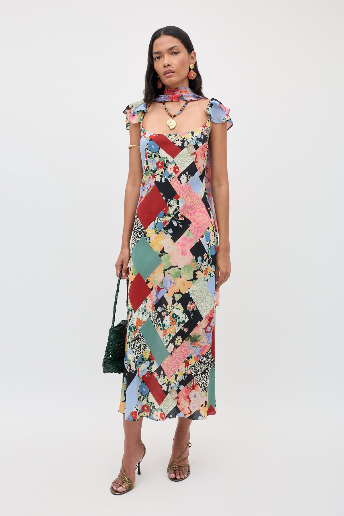 A woman wearing the Adele - Olivia Patchwork Multi midi dress with ruffled sleeves, paired with a matching scarf and sandals, holding a dark green handbag and standing against a plain white background.
