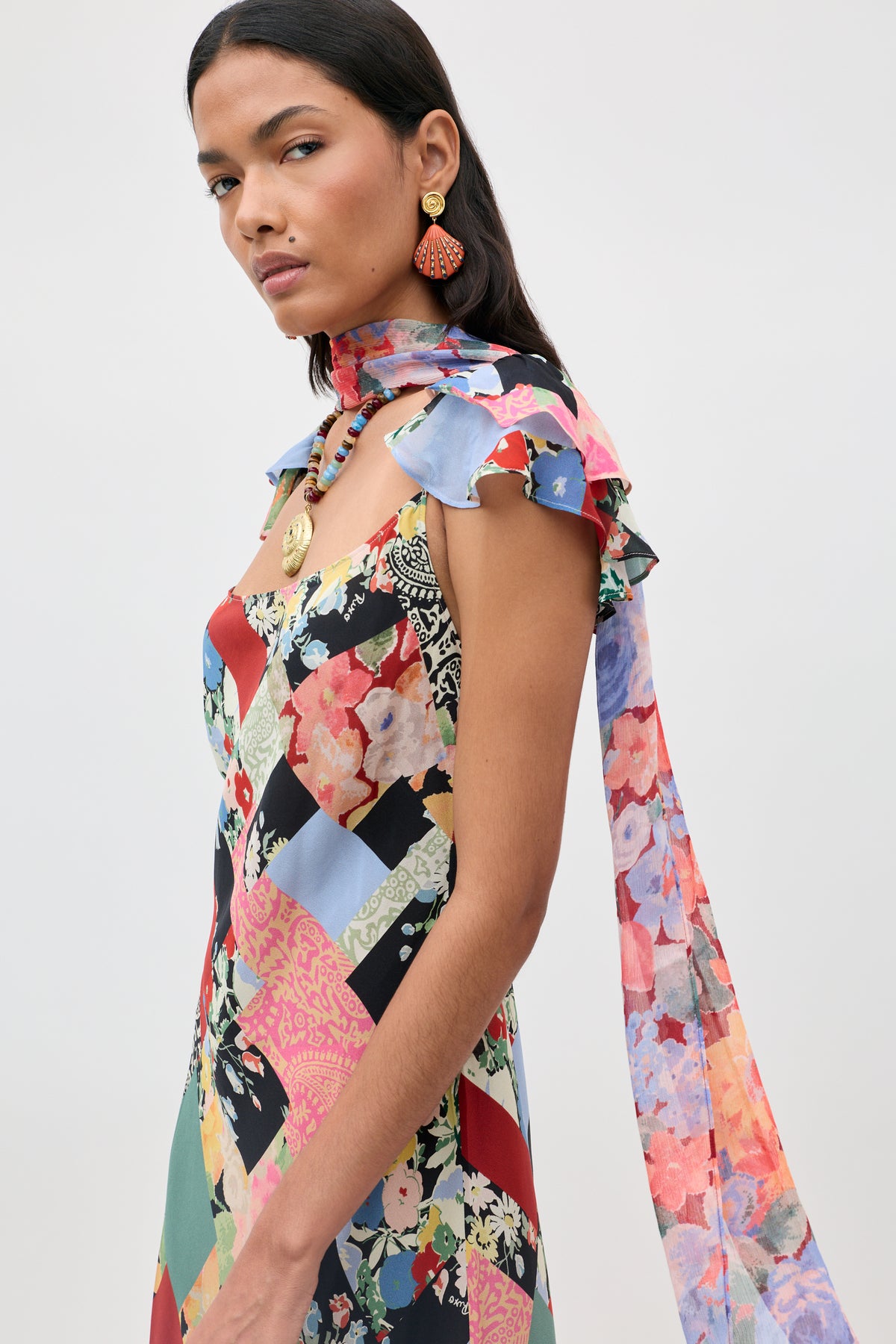 A woman wearing the Adele - Olivia Patchwork Multi dress with a long matching scarf and statement earrings, looking sideways against a plain light background.