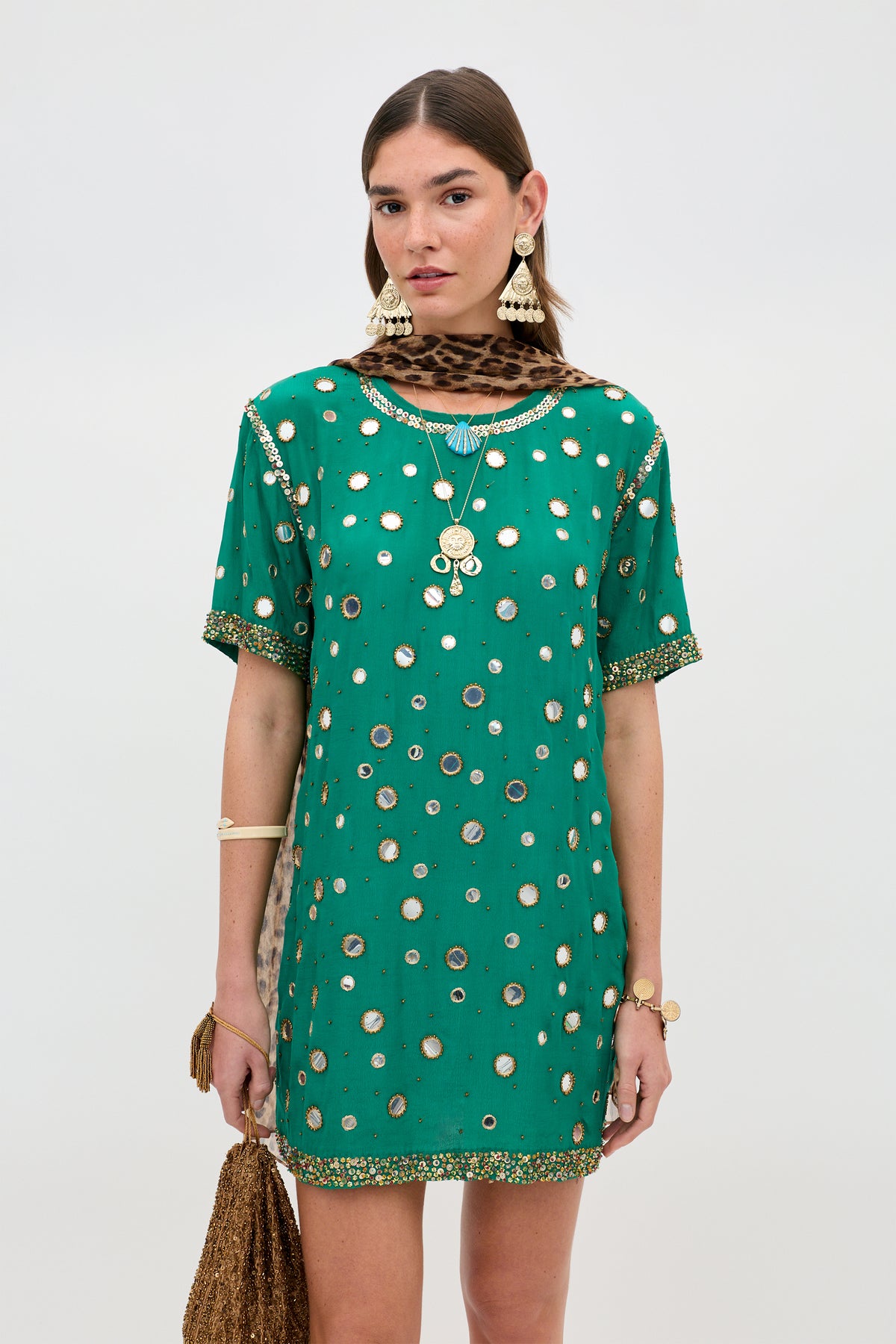 A woman stands against a plain background, wearing the Cruz - Lume Embellishment Dark Green short dress paired with gold jewelry, statement earrings, and a leopard print scarf.