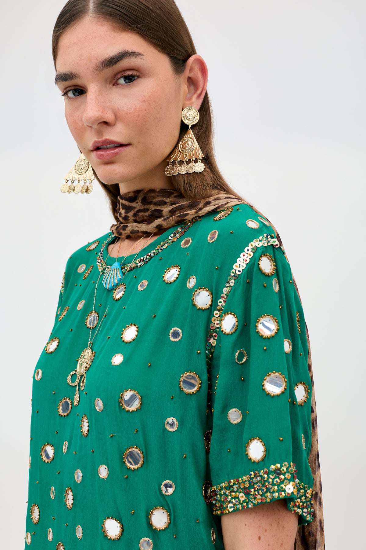 A person wearing the Cruz - Lume Embellishment Dark Green dress, adorned with mirrors and sequins, gold statement earrings, a necklace, and a brown leopard print scarf poses against a plain background.