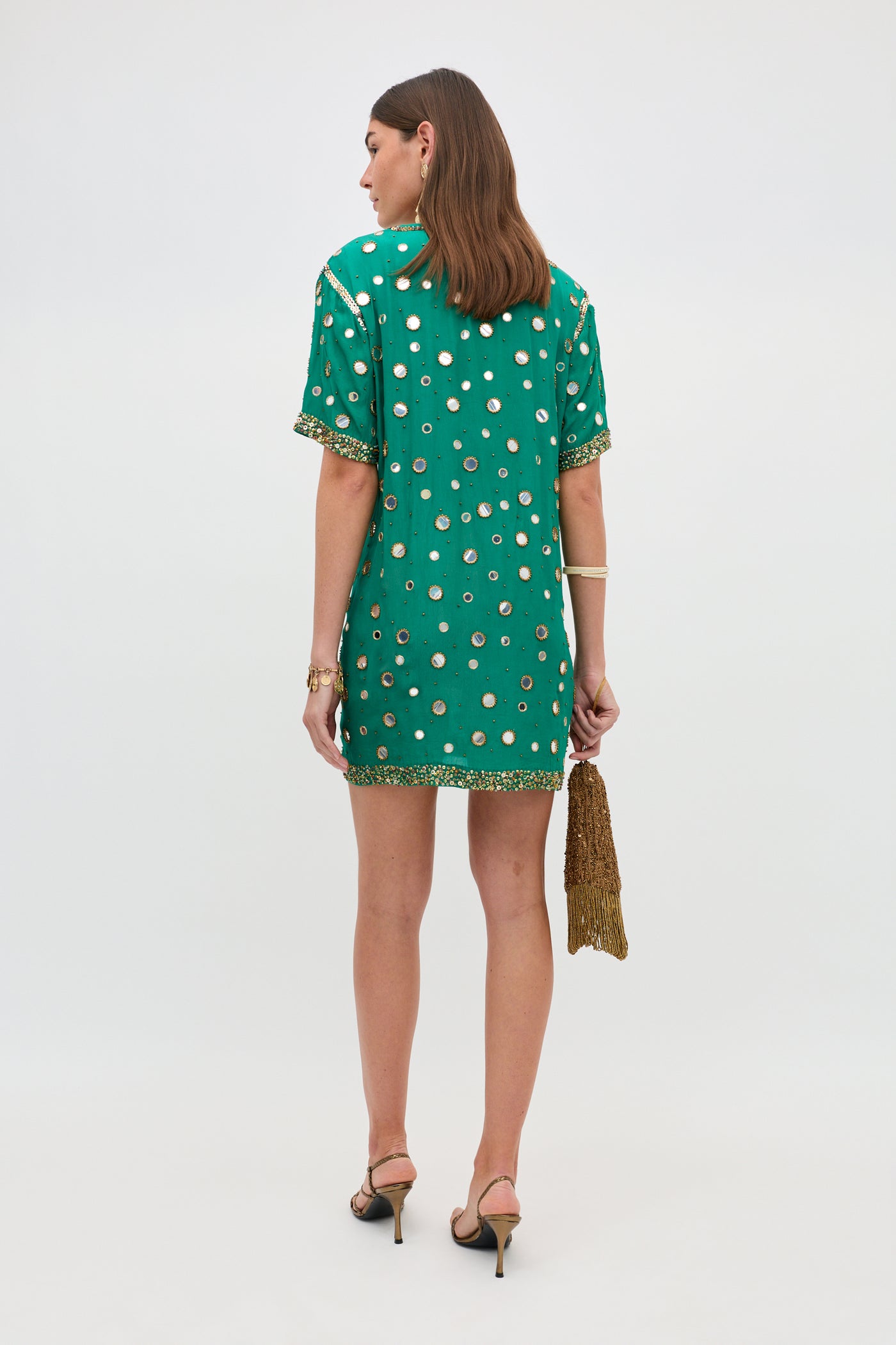 A woman stands facing away, wearing the Cruz - Lume Embellishment Dark Green dress, a short green dress adorned with gold circle accents. She pairs it with black heels and a fringed gold clutch, and her medium-length brown hair is styled down. The background is plain white.