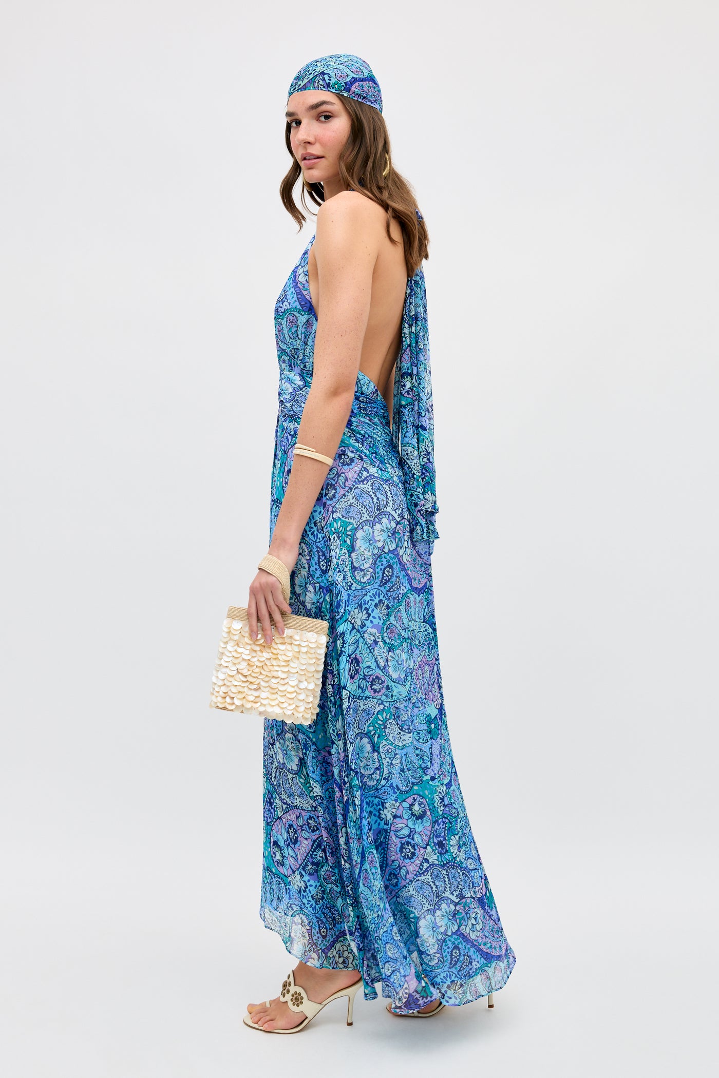A woman stands sideways against a plain background, wearing the Alaina - Paisley Floral Blue dress with an open back, a matching headscarf, beige heeled sandals, gold jewelry, and carrying a woven handbag.