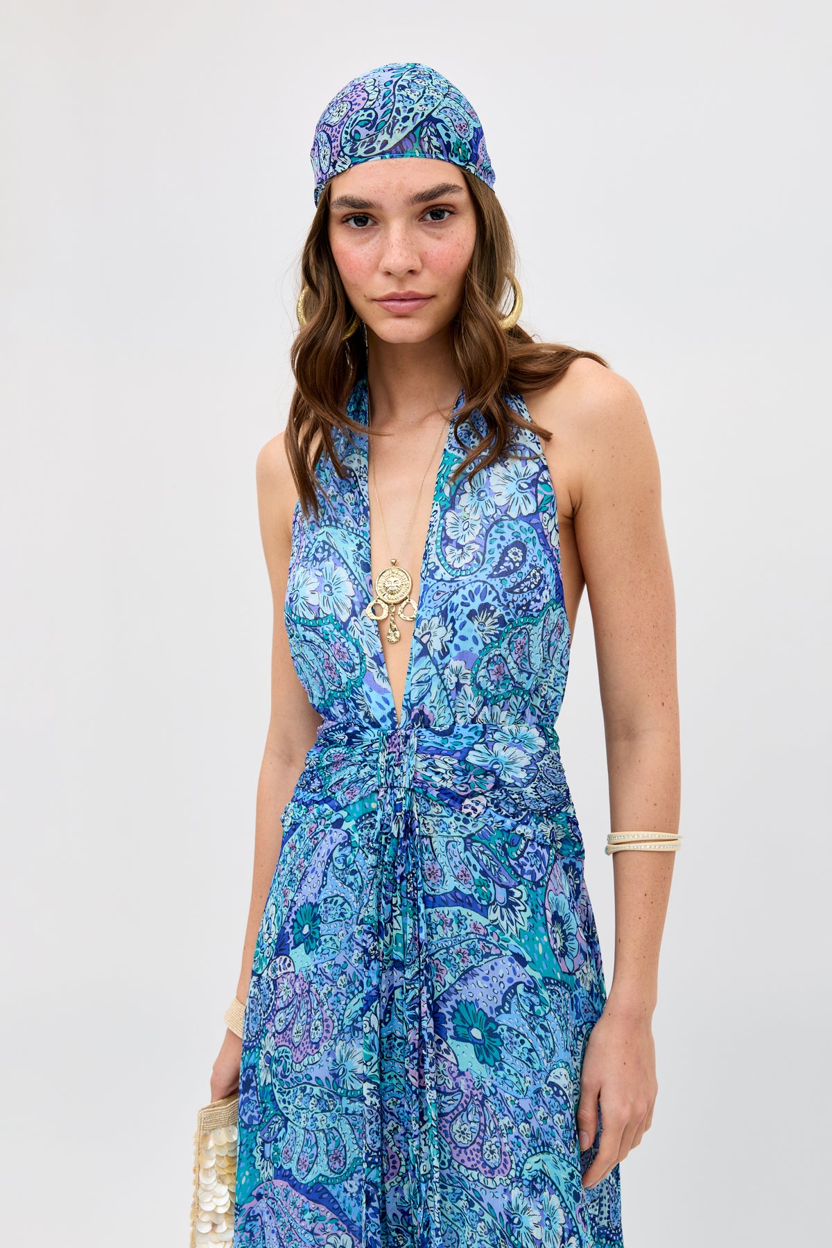 Wearing the Alaina - Paisley Floral Blue halter dress and matching headscarf, a woman accessorized with gold jewelry and bracelets stands against a plain light background, holding a small clutch.