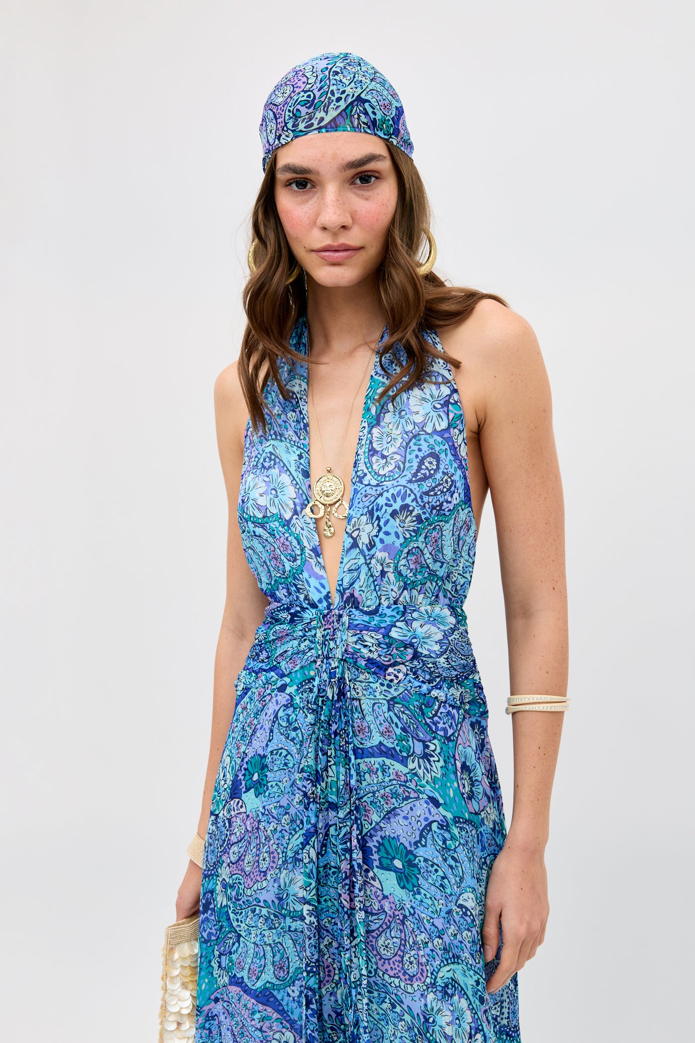 Wearing the Alaina - Paisley Floral Blue halter dress and matching headscarf, a woman accessorized with gold jewelry and bracelets stands against a plain light background, holding a small clutch.