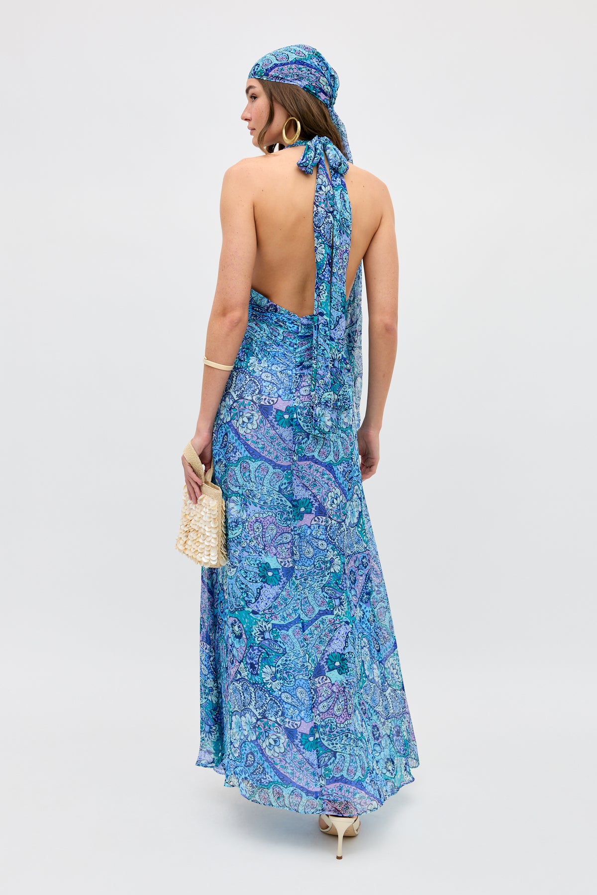 Wearing the Alaina - Paisley Floral Blue backless halter dress and matching headscarf, a woman stands with her back to the camera, holding a woven beige purse and accessorizing with hoop earrings, a bracelet, and light heels.