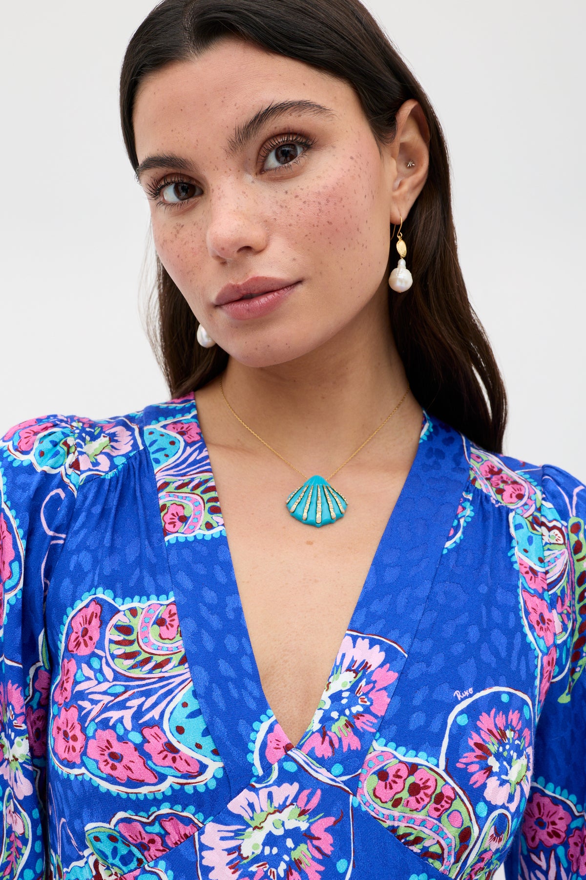 A woman with long dark hair and freckles wears a blue paisley dress and the Leanira - Turquoise necklace, featuring a gold chain and turquoise seashell pendant. She gazes calmly at the camera.