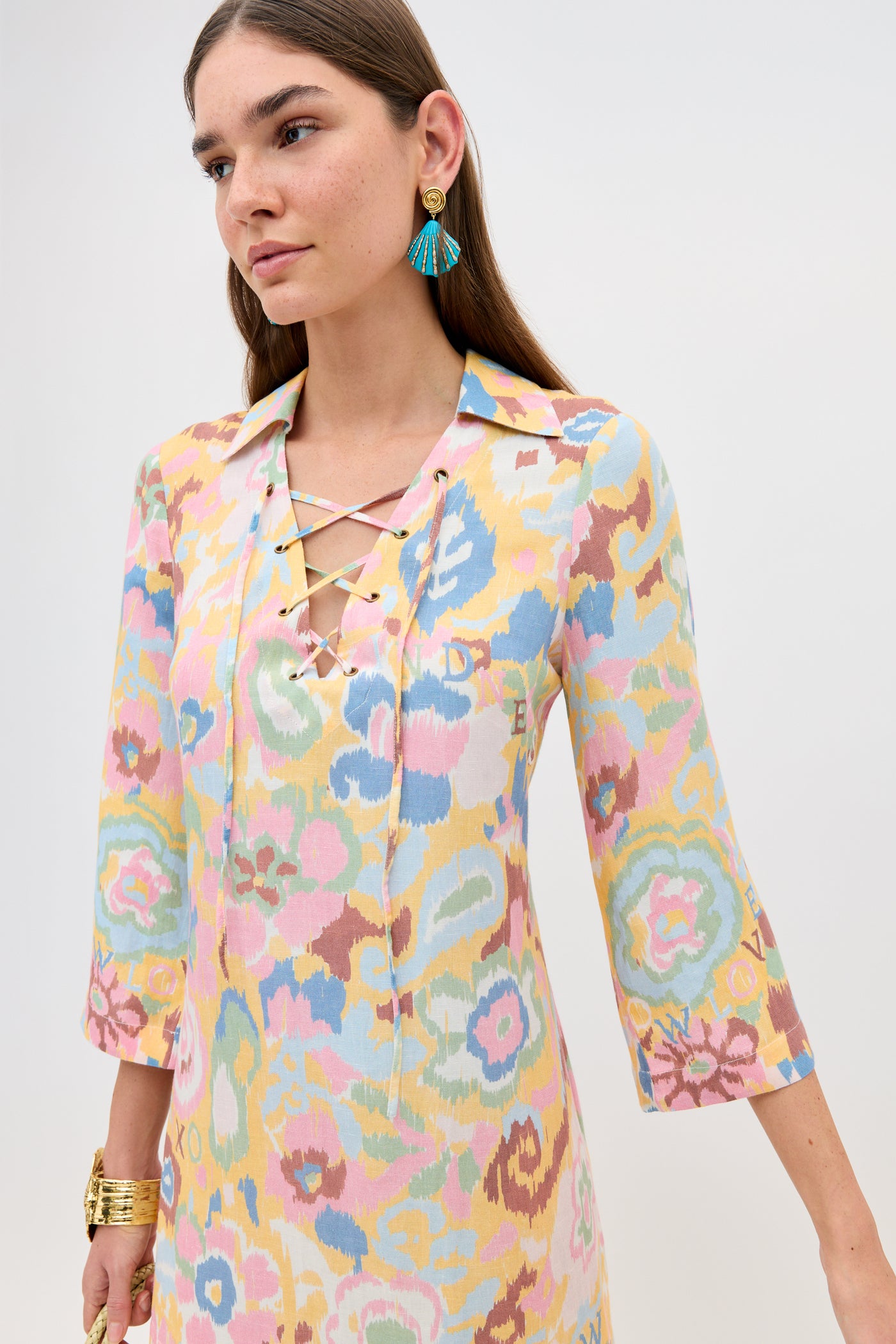 A woman in the Lucien - Ikat Bloom Yellow dress with a lace-up neckline and three-quarter sleeves, straight brown hair, gold bracelets, and blue tassel earrings stands against a plain light background.