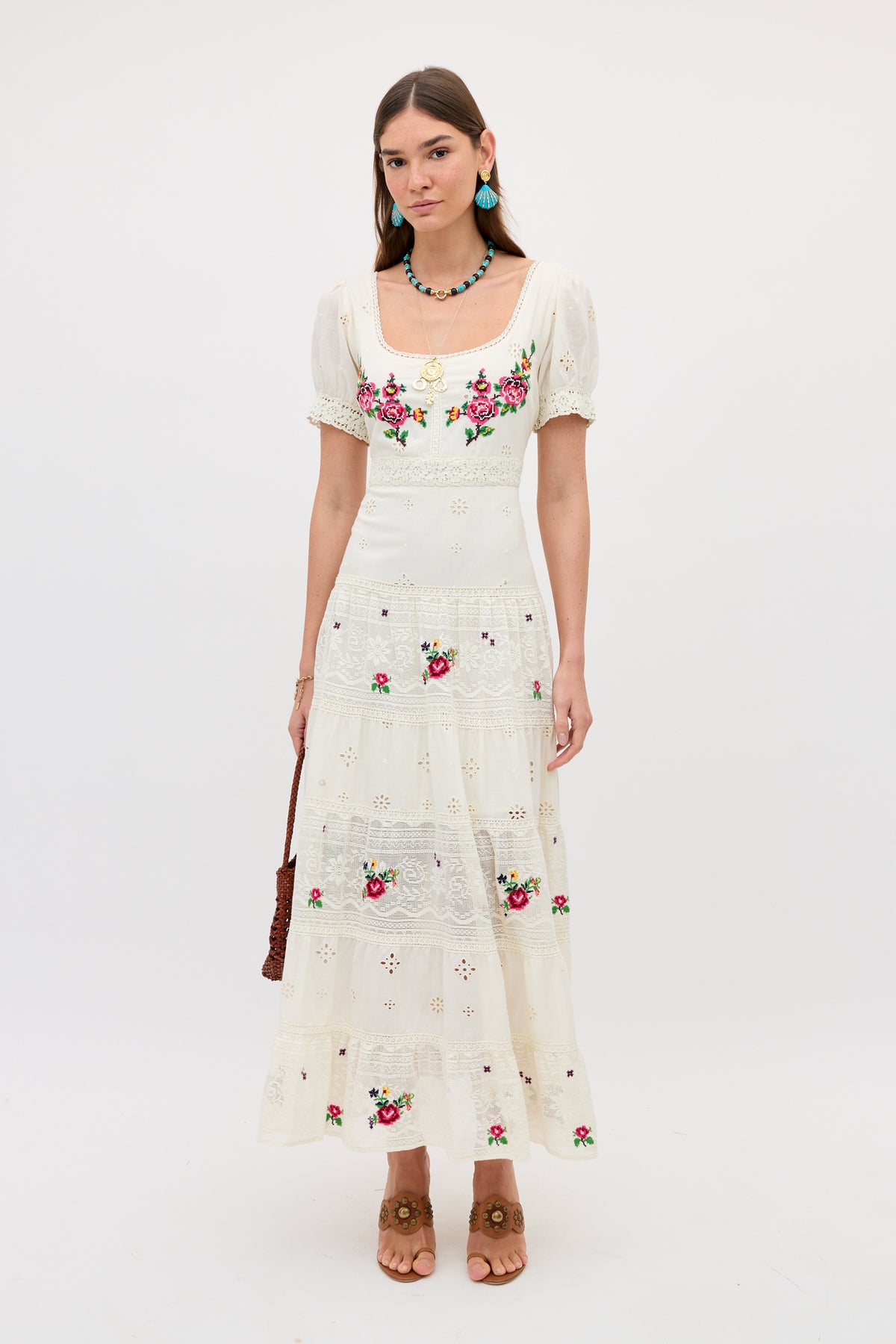 A woman wears the Cady - Spring Rose Embroidery Cream dress, featuring floral embroidery and lace details. She completes the look with turquoise earrings, a beaded necklace, a brown purse, and tan heeled sandals.