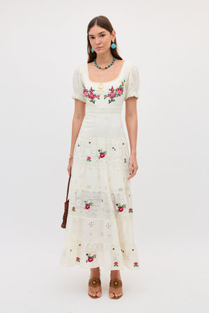 A woman wears the Cady - Spring Rose Embroidery Cream dress, featuring floral embroidery and lace details. She completes the look with turquoise earrings, a beaded necklace, a brown purse, and tan heeled sandals.