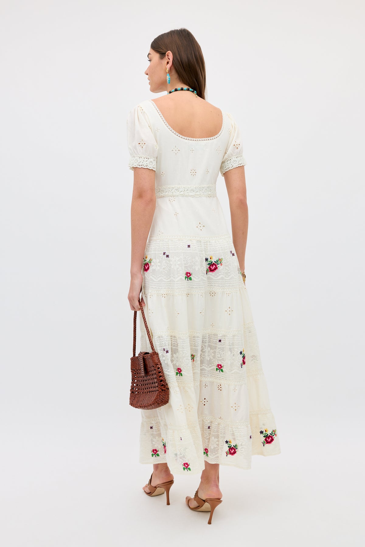 A woman wears the Cady - Spring Rose Embroidery Cream dress, ankle-length with short sleeves and open back. She faces away, carrying a brown woven handbag and wearing tan heeled sandals, blue earrings, and a necklace.