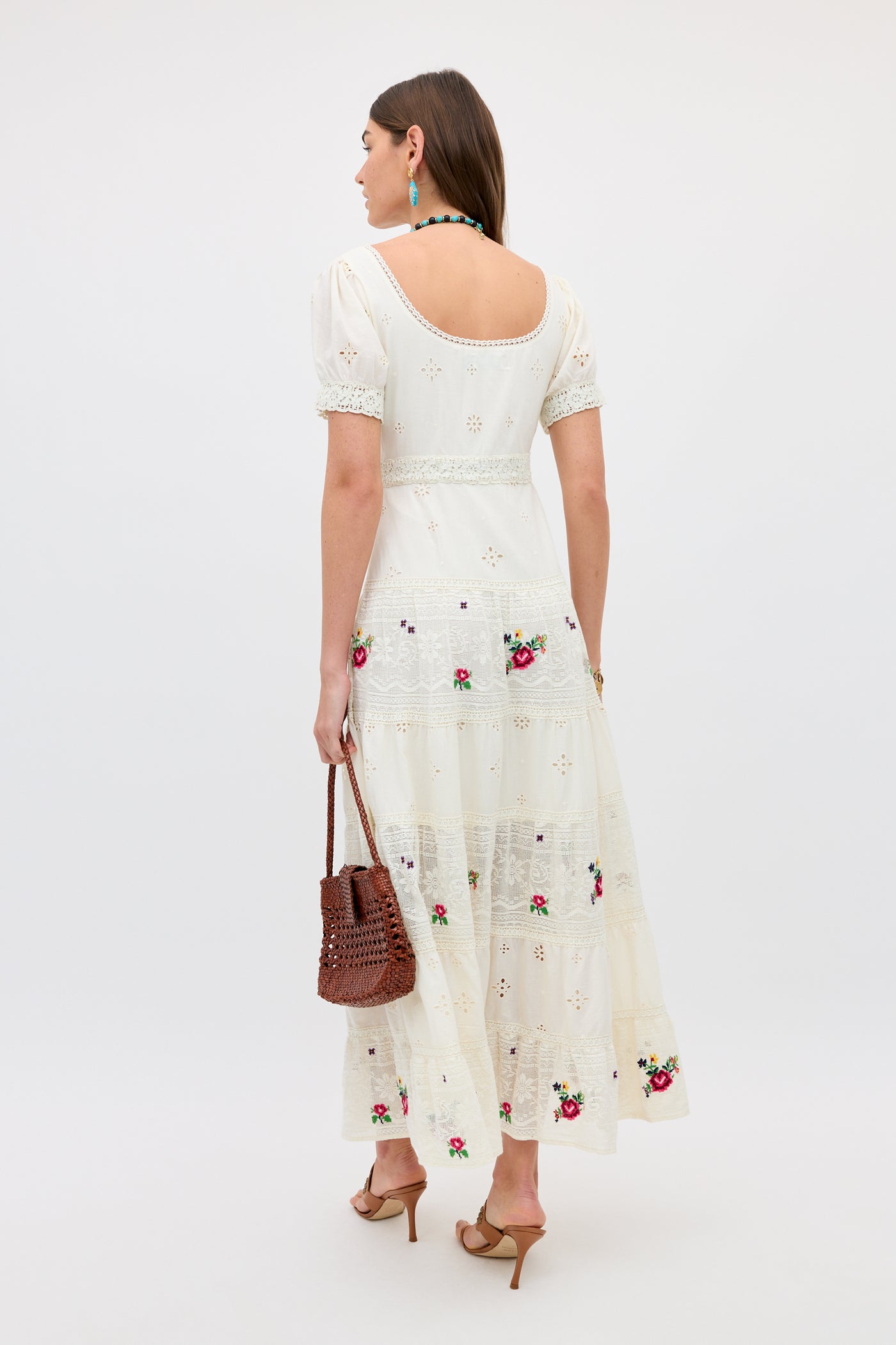 A woman wears the Cady - Spring Rose Embroidery Cream dress, ankle-length with short sleeves and open back. She faces away, carrying a brown woven handbag and wearing tan heeled sandals, blue earrings, and a necklace.