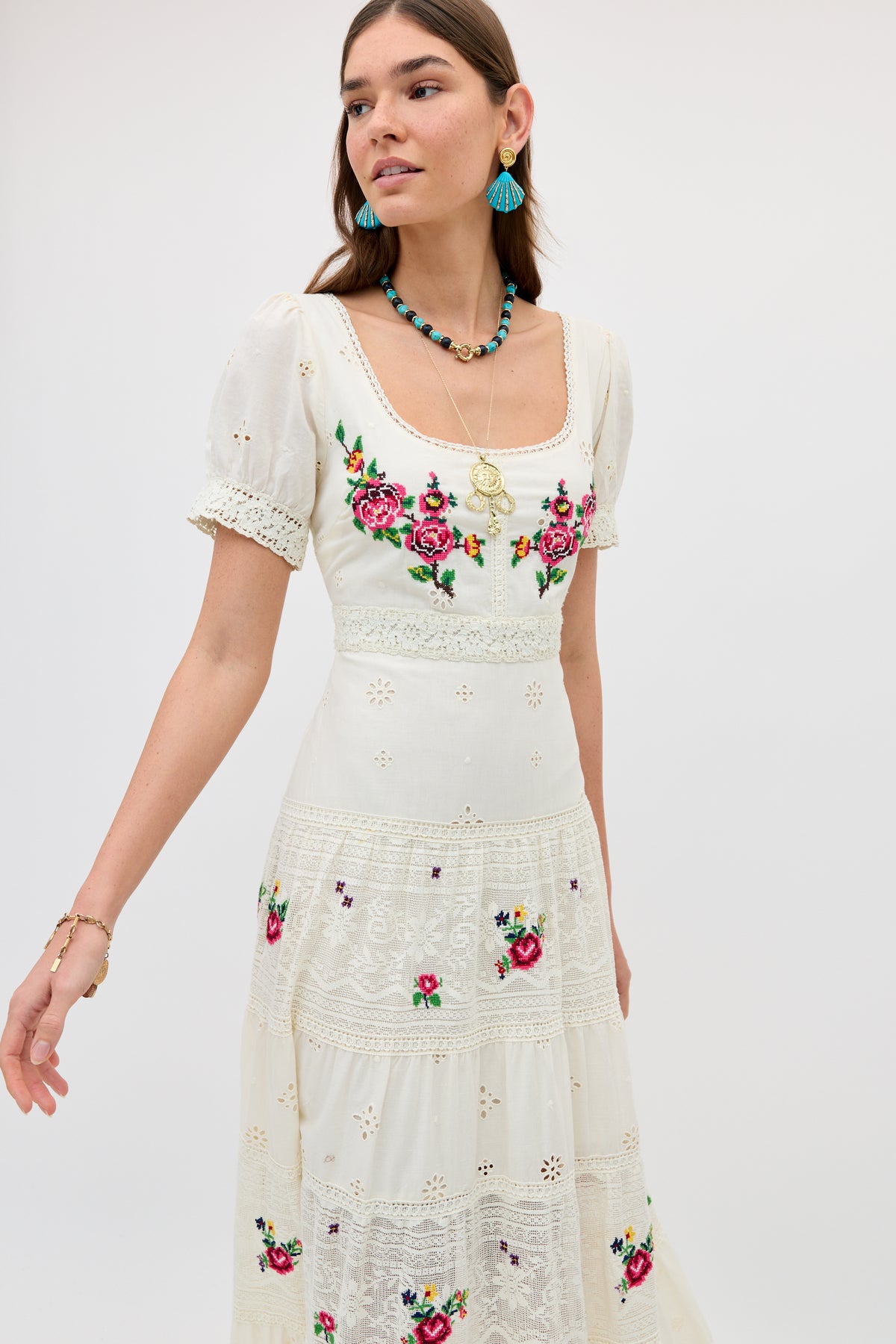 A woman wears the Cady - Spring Rose Embroidery Cream dress, featuring white short sleeves, colorful floral embroidery, and lace details. She accessorizes with blue earrings, layered necklaces, and a bracelet against a light background.