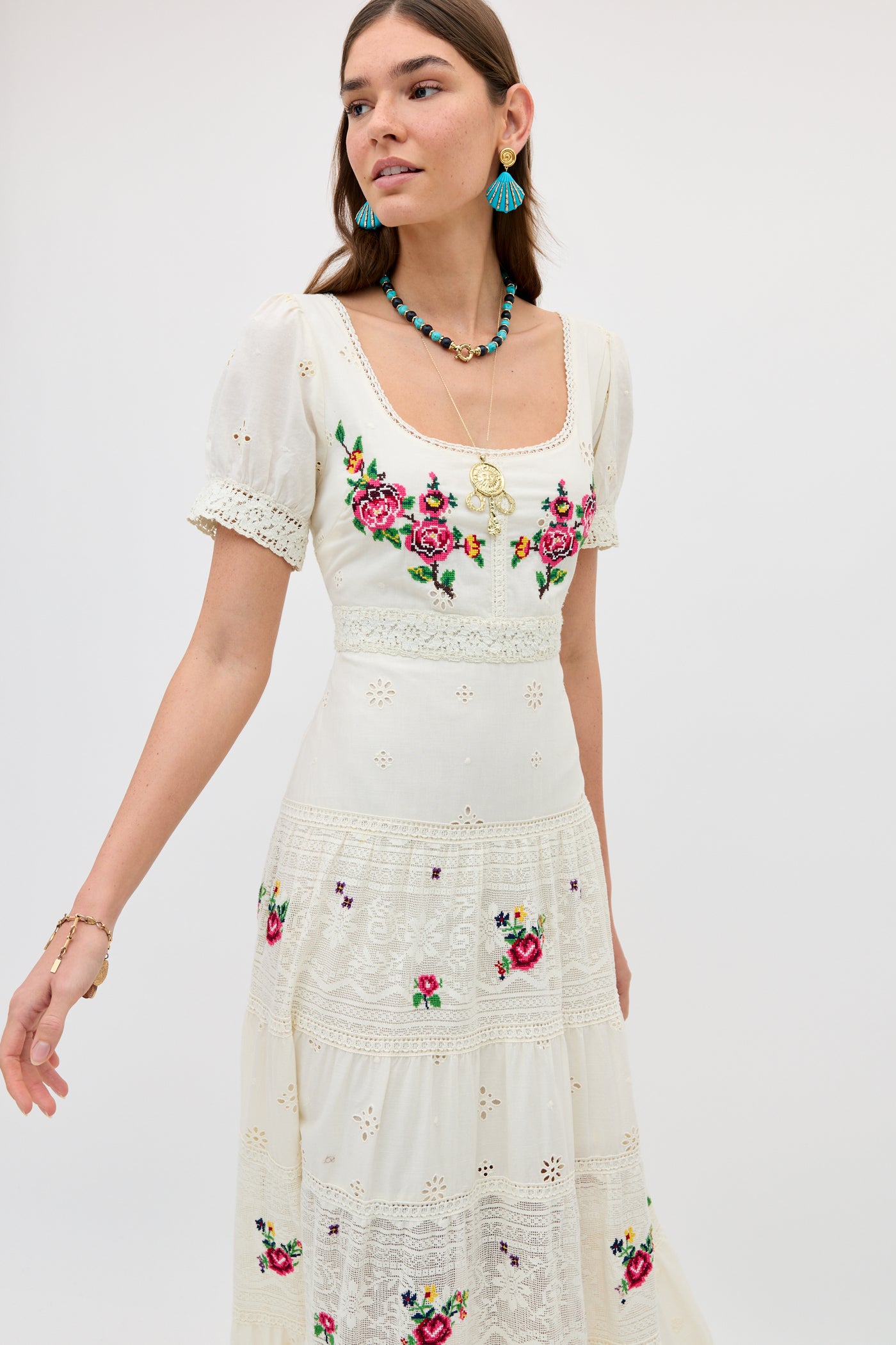 A woman wears the Cady - Spring Rose Embroidery Cream dress, featuring white short sleeves, colorful floral embroidery, and lace details. She accessorizes with blue earrings, layered necklaces, and a bracelet against a light background.