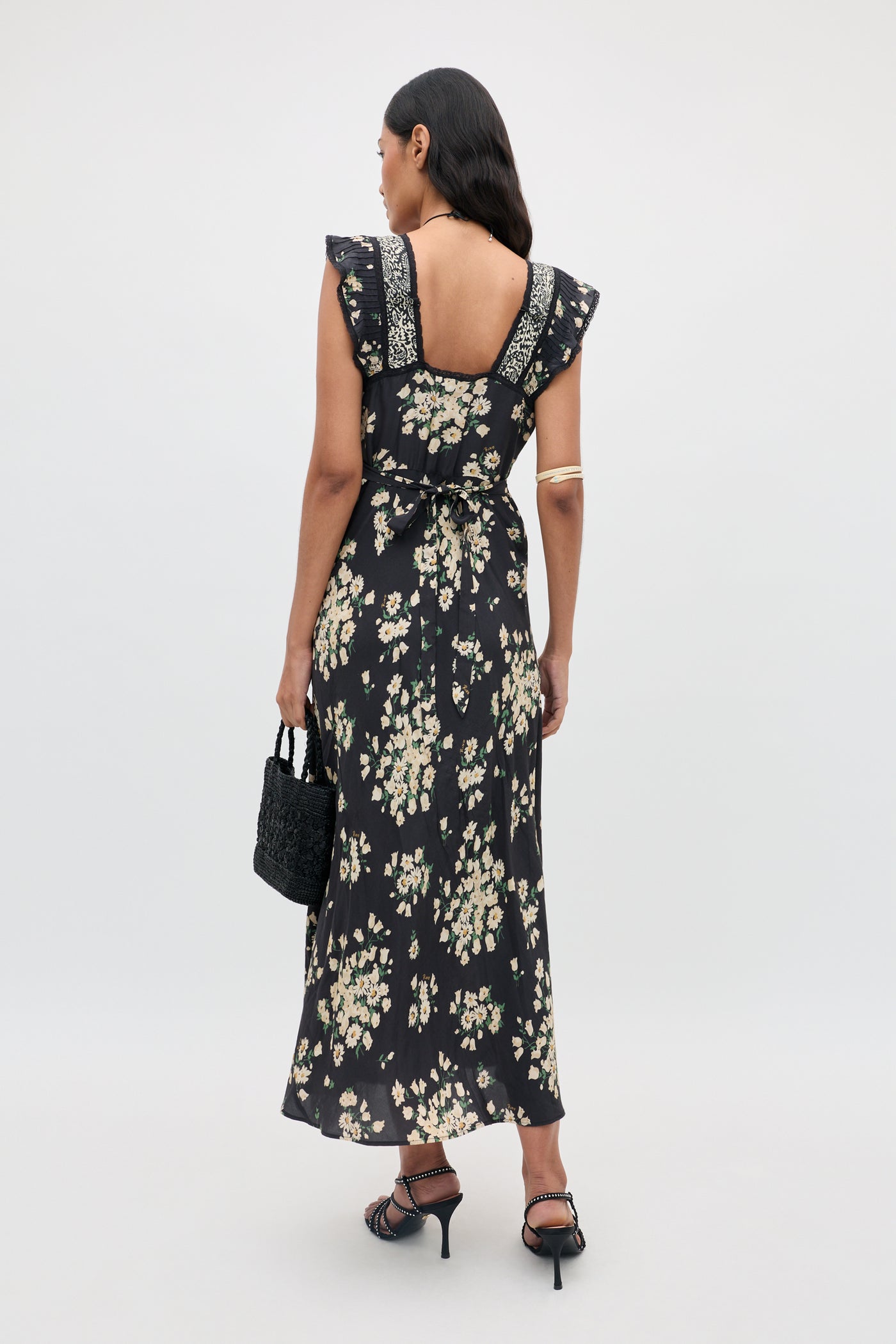 A woman stands with her back to the camera, wearing the Bailee - Amelia Gremlin Mix Black dress featuring ruffle sleeves. She completes her look with a black handbag, black heels, and a gold arm cuff against a plain light grey background.