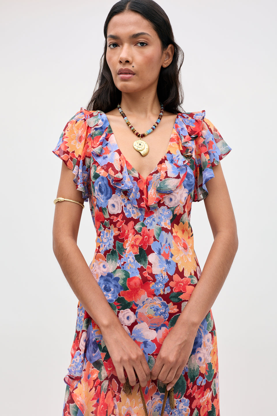 A woman stands confidently against a plain background wearing the Meadow - Chelsea Floral Red dress, featuring ruffled sleeves and a vibrant floral pattern. She has long dark hair, wears a necklace and a gold armband, and gazes directly at the camera.