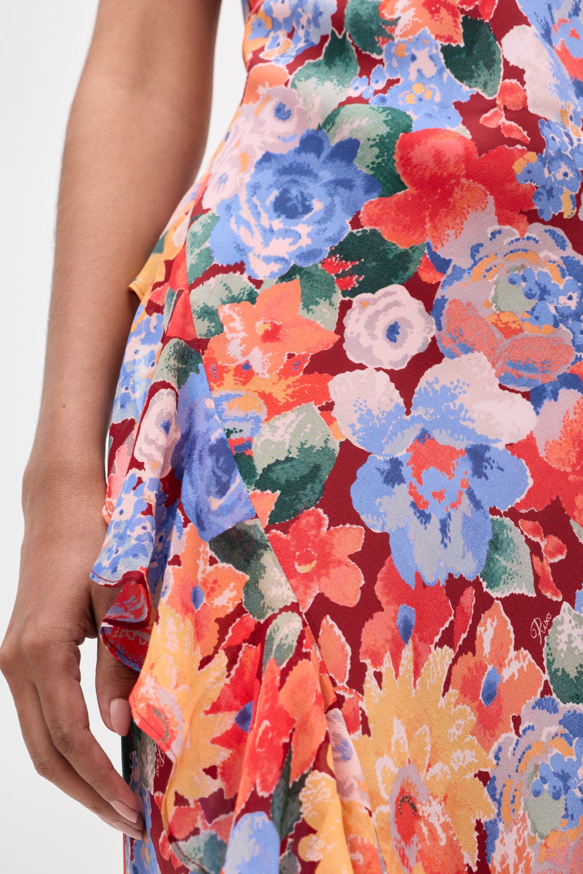A person wearing the "Meadow - Chelsea Floral Red" dress, featuring a vibrant floral pattern in red, blue, orange, and yellow; only their hand and part of their body are visible.