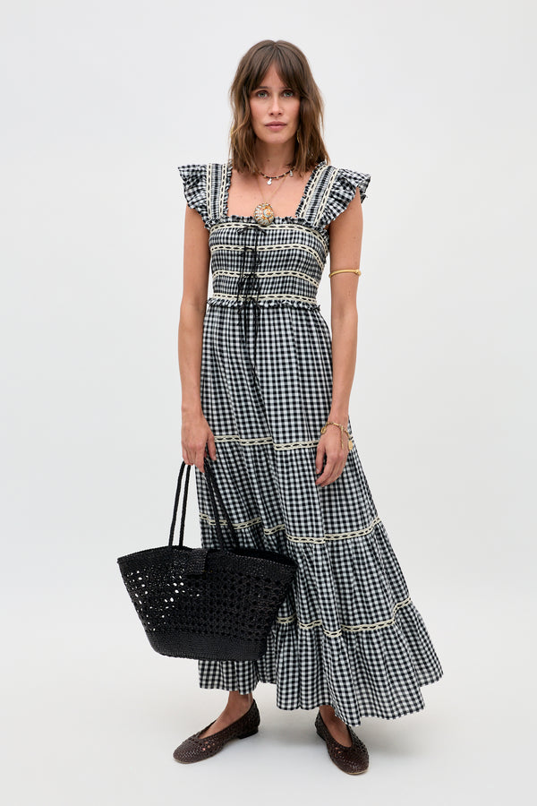 A woman stands against a plain background wearing the Chandra - Beth Check Black maxi dress with ruffled sleeves. She completes the look with a black woven tote, brown flats, layered necklaces, and an arm cuff.